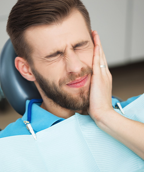 Emergency services for tooth injuries, loose teeth, pain, swelling, dental abscess, lost crowns, wisdom tooth pain, and more is available at our dental office in West Bloomfield, Michigan.  In this image, a man in a dentist's chair holds his cheek in pain.