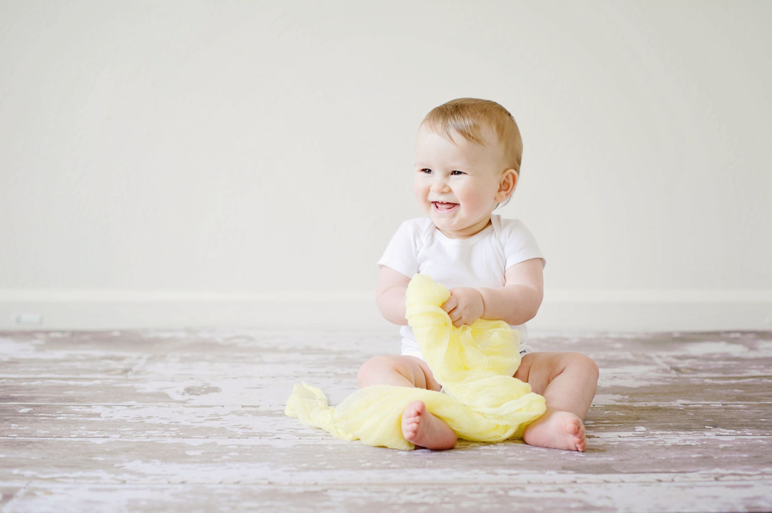 An infant sits on a wooden floor while holding a yellow dress and smiling, revealing four small teeth. This dental blog features 16 facts about baby teeth for new parents.