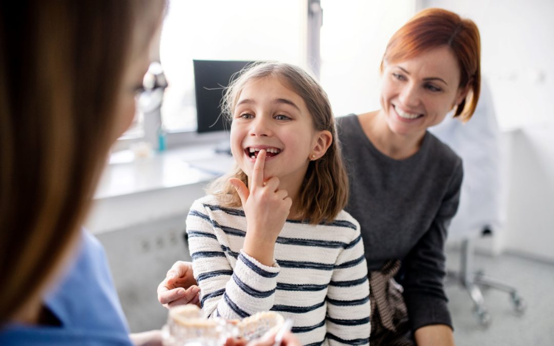 Our family dentistry in West Bloomfield, Michigan offers pedodontics. In this image, a young girl is smiling as she shows a dental professional her missing tooth as the girl's mother looks on.