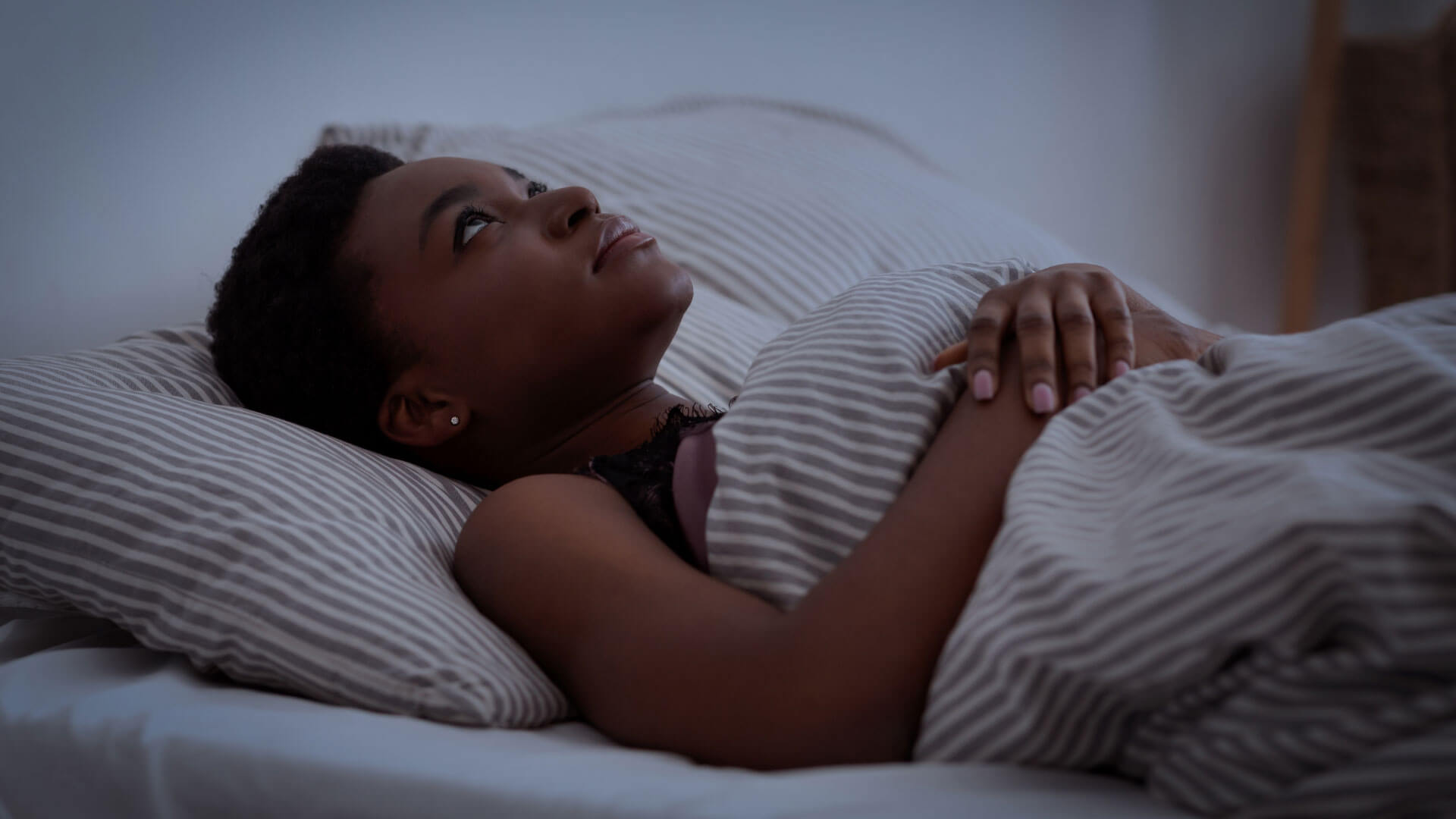 A woman lying in bed under striped blankets, looking upward with a contemplative expression.