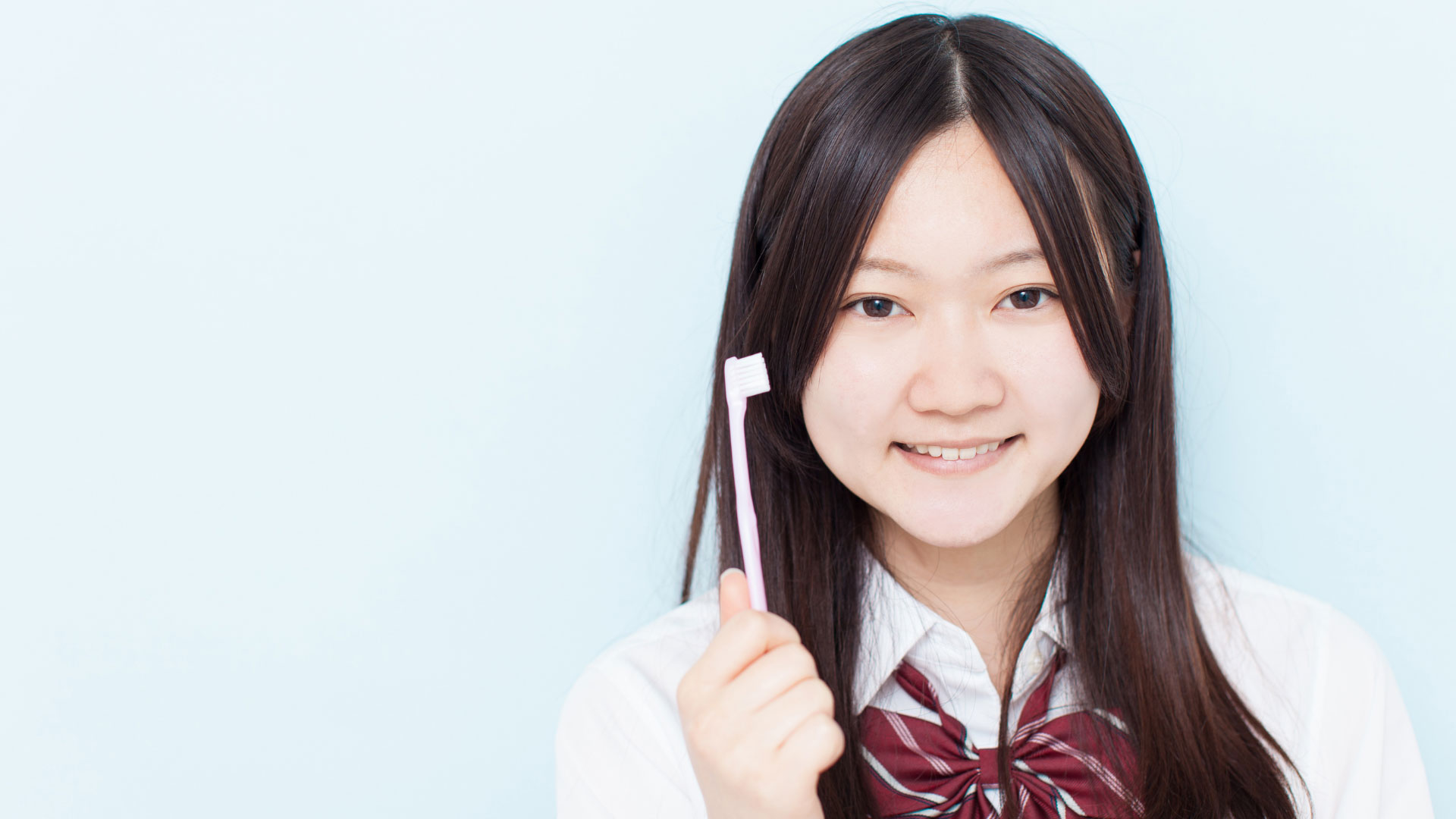 A young student smiles at the camera, holding up a light pink toothbrush.